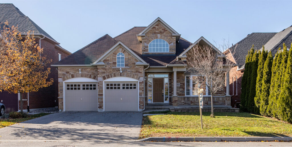 A two-story brick house with a pitched shingle roof and a double garage featuring arched windows above the garage doors. The house number "602" is visible near the centered front door. The property includes a front yard with a tree bearing autumn leaves on the left and a leafless tree on the right. The driveway leads to the garage, with a sidewalk and some grass in front. Similar neighboring houses are visible on either side, and tall green shrubs line the right edge of the yard.